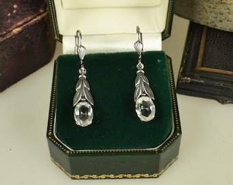 May include: A pair of silver drop earrings featuring oval-cut clear stones suspended from a metal leaf-shaped design. The earrings have lever-back closures and are presented in a dark green velvet-lined box with the text The Royal Collection.