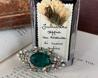 Könnte beinhalten: Eine vintage Brosche aus grünem Glas mit einer goldenen Fassung und klaren Strasssteinen. Die Brosche ist blattförmig und hat ein filigranes Design. Die Brosche liegt auf einem Buch mit dem sichtbaren Text "Jucluastro seppia cou esseusa di cuoio antico".