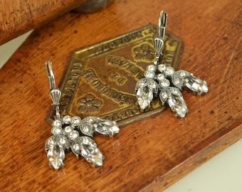 May include: A pair of silver-toned leverback earrings featuring a fan-shaped design. Each earring is adorned with multiple clear round rhinestones and three marquise-cut clear crystals set in a textured, darkened metal frame with a metallic finish.