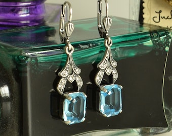 May include: A pair of drop earrings featuring rectangular light blue faceted stones suspended from silver-toned metal settings. The upper portion of each earring displays a decorative filigree design accented with small clear stones and lever-back closures.