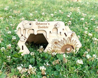 May include: A light-colored wooden bee house featuring a hexagonal entrance, engraved sunflower and honeycomb patterns, and decorative bee cutouts. The top edge has a dripping honey design with the text "Rebecca's BeeHide" engraved in the center.