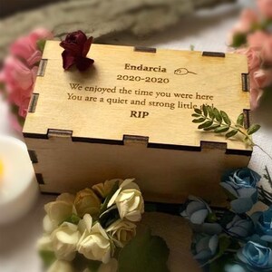 K&ouml;nnte beinhalten: Eine hellfarbene Holzkiste mit einer lasergravierten Inschrift, umgeben von Blumen. Der Text lautet: "Endarcia 2020-2020, Wir haben die Zeit genossen, die du hier warst, Du bist ein stilles und starkes kleines, RIP."