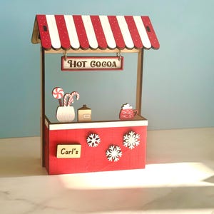 May include: A miniature wooden hot cocoa stand with a red and white striped awning. The stand has a sign saying "Hot Cocoa", snowflakes, and decorative elements like candy canes and a cocoa mug. The sign reads "Carl's".