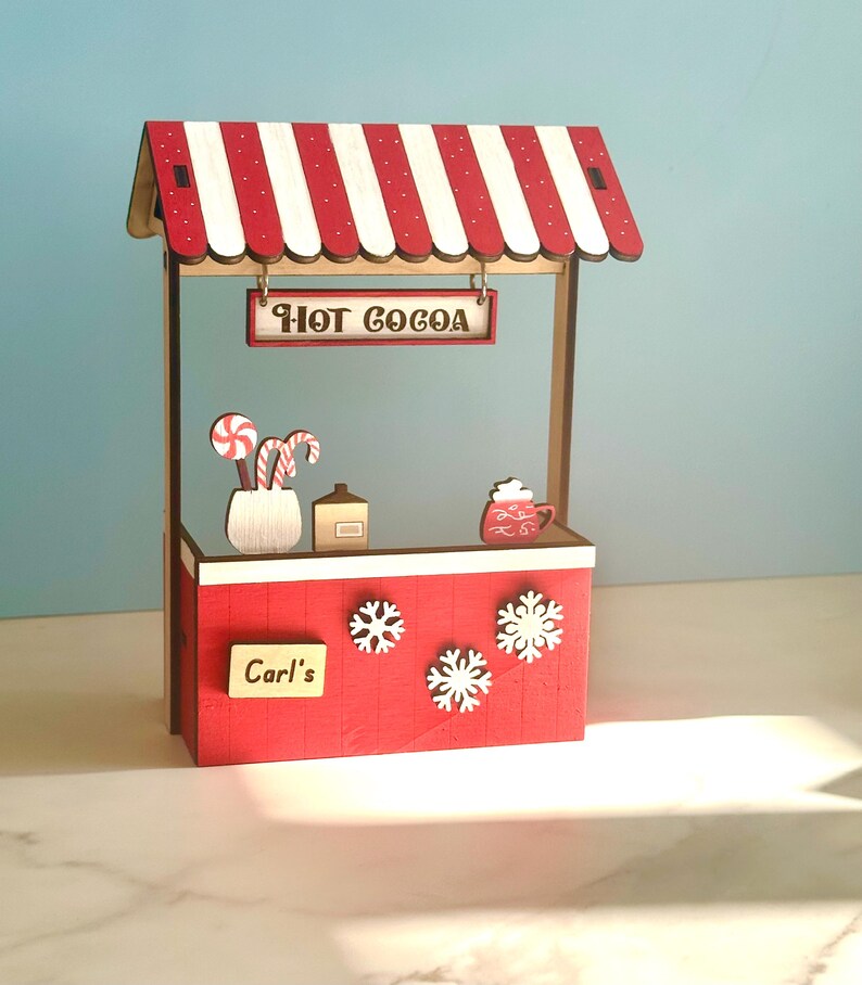 May include: A miniature wooden hot cocoa stand with a red and white striped awning. The stand has a sign saying "Hot Cocoa", snowflakes, and decorative elements like candy canes and a cocoa mug. The sign reads "Carl's".