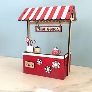 May include: A miniature red and white striped hot cocoa stand with a sign that reads "HOT COCOA". The stand has a red base with snowflakes and the name "Carl's". Decorative candy canes and a mug of cocoa are visible.