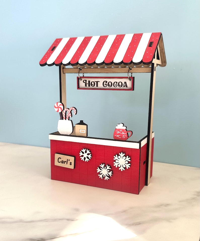 May include: A miniature red and white striped hot cocoa stand with a sign that reads "HOT COCOA". The stand has a red base with snowflakes and the name "Carl's". Decorative candy canes and a mug of cocoa are visible.