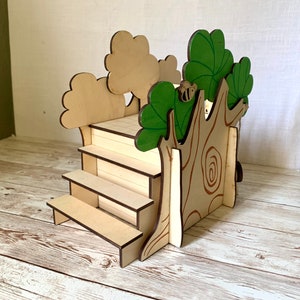 Puede incluir: Expositor de madera con tem&aacute;tica de casa en el &aacute;rbol, con escalones y una plataforma. El tronco del &aacute;rbol presenta detalles de vetas de madera y una peque&ntilde;a abeja. Las hojas est&aacute;n pintadas de verde y los &aacute;rboles son de color marr&oacute;n claro. El expositor mide unos 15 cm de alto.