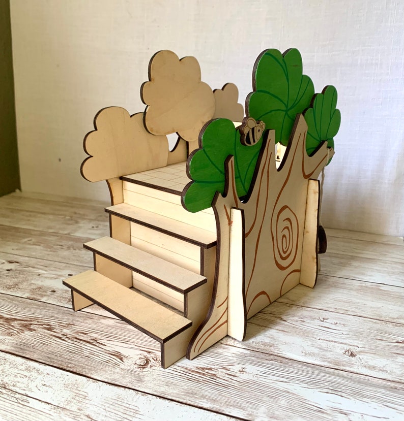 Puede incluir: Expositor de madera con tem&aacute;tica de casa en el &aacute;rbol, con escalones y una plataforma. El tronco del &aacute;rbol presenta detalles de vetas de madera y una peque&ntilde;a abeja. Las hojas est&aacute;n pintadas de verde y los &aacute;rboles son de color marr&oacute;n claro. El expositor mide unos 15 cm de alto.
