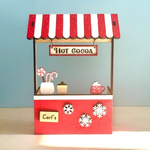 May include: A miniature red and white striped hot cocoa stand with the text "HOT COCOA" on a sign. The stand features snowflake decorations, a sign that says "Carl's", and decorative candy canes.