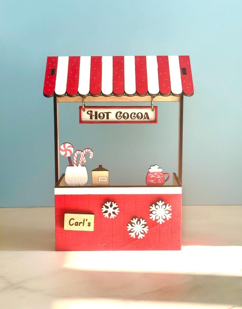 May include: A miniature red and white striped hot cocoa stand with the text "HOT COCOA" on a sign. The stand features snowflake decorations, a sign that says "Carl's", and decorative candy canes.