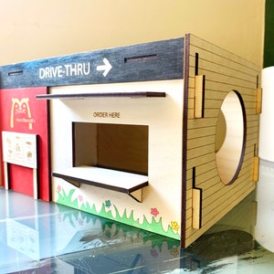 May include: A wooden model of a drive-thru restaurant with a red and white facade, a drive-thru window, and a sign that says "DRIVE-THRU". The model is painted with brown, black, and white paint and has a small green lawn in front of it.