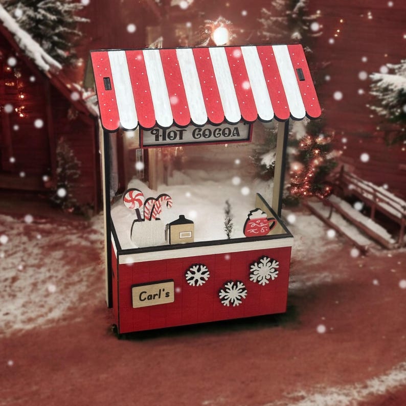 May include: A miniature red and white striped hot cocoa stall with the text "Hot Cocoa" above the opening. The stall has snowflake decorations and the name "Carl's" on the side. Candy canes and a mug are visible inside.