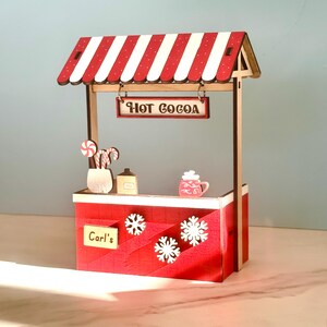 May include: A miniature wooden hot cocoa stand with a red and white striped awning. The stand has a sign saying "HOT COCOA", snowflakes, and "Carl's". Decorative candy canes, a mug, and a small house are also visible.