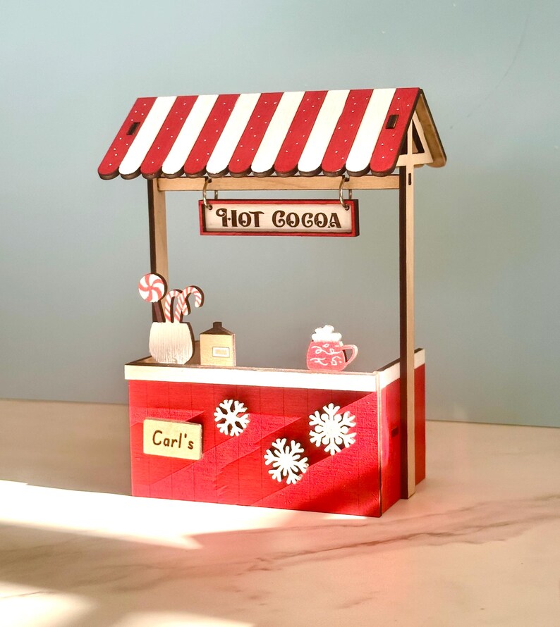 May include: A miniature wooden hot cocoa stand with a red and white striped awning. The stand has a sign saying "HOT COCOA", snowflakes, and "Carl's". Decorative candy canes, a mug, and a small house are also visible.