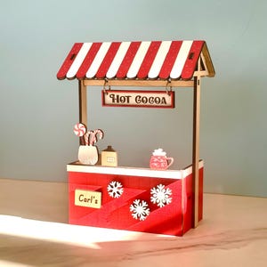 May include: A miniature wooden hot cocoa stand with a red and white striped awning. The stand features a sign that reads "HOT COCOA", snowflake cutouts, and a sign that reads "Carl's". Decorative items include candy canes, a mug, and a bottle.