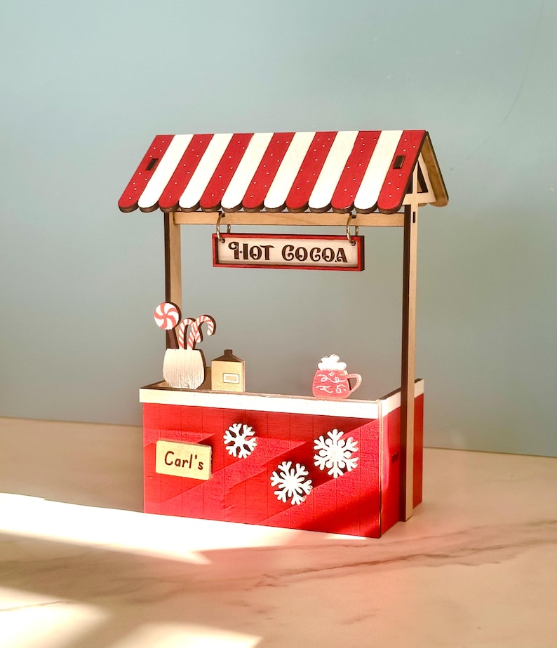 May include: A miniature wooden hot cocoa stand with a red and white striped awning. The stand features a sign that reads "HOT COCOA", snowflake cutouts, and a sign that reads "Carl's". Decorative items include candy canes, a mug, and a bottle.