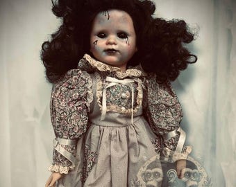 May include: A porcelain doll with dark curly hair and a painted face. The doll is wearing a gray and white dress with floral accents and lace trim. The doll is holding a card with a picture of two monkeys and a smaller doll.