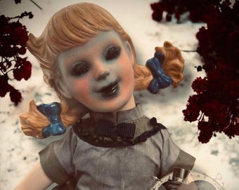 May include: A porcelain doll with blue eyes and a wide smile, wearing a brown dress and blue ribbons in her hair. The doll is holding a circular image of three monkeys with a smaller doll in the center.