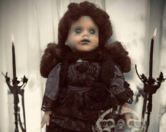 May include: A porcelain doll with dark brown hair, wearing a black and purple dress with lace trim, stands on a wooden surface with candles and flowers. The doll has a pale complexion and blue eyes.