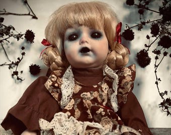 May include: A porcelain doll with blonde hair in braids, wearing a brown dress with a floral pattern and lace trim. The doll has blue eyes and a painted expression. The doll is sitting on a wooden surface with a background of dried flowers.
