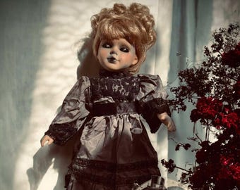 May include: A porcelain doll with a gray dress and black lace trim. The doll has blonde hair styled in a bun and painted-on blue eyes. The doll is standing on a wooden surface with a vintage-style illustration of three monkeys behind it.