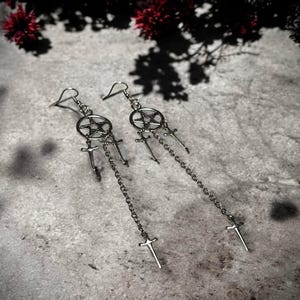 May include: A pair of silver earrings with a pentagram charm and dangling swords. The earrings are on a gray background.