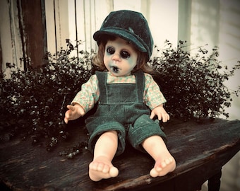 May include: A spooky doll with dark eye makeup and a black cap. The doll wears a green corduroy overall and a patterned shirt. It sits on a weathered wooden surface, with dark foliage in the background.