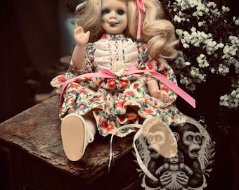 May include: A vintage porcelain doll with blonde hair and a floral dress. The doll has a pale complexion with dark eye makeup and a pink ribbon in her hair. A tag with a monkey illustration hangs nearby.