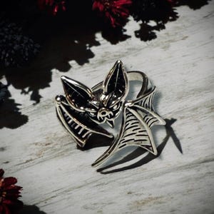 Może przedstawiać: Srebrny pierścionek z detalicznym wzorem nietoperza. Skrzydła nietoperza są rozłożone, a nietoperz patrzy wprost z widocznymi kłami.