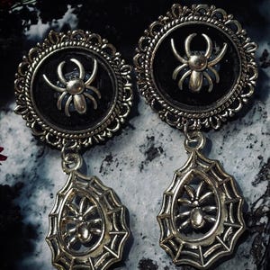 May include: A pair of silver dangle earrings featuring a black spider design. The top of each earring is a round, silver frame with a black background and a silver spider in the centre. The bottom of each earring is a silver spiderweb design with a silver spider in the centre.