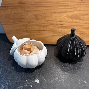 Puede incluir: Dos recipientes con forma de ajo, uno blanco y otro negro, sobre una superficie oscura. El recipiente blanco está abierto, lleno de dientes de ajo. Una tabla de cortar de madera en el fondo.
