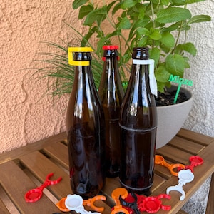Könnte beinhalten: Drei braune Glasflaschen mit farbigen Flaschenverschluss-Haltern. Die Verschlüsse sind gelb, rot und weiß. Mehrere Flaschenverschluss-Halter in verschiedenen Farben liegen auf einer Holzoberfläche. Eine Topfpflanze befindet sich im Hintergrund.