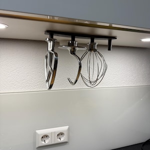 Könnte beinhalten: Drei silberne Küchenmixer-Aufsätze, die an einem Metallgestell hängen, das unter einem Schrank montiert ist. Die Aufsätze umfassen einen Knethaken, einen Schneebesen und einen Flachrührer.