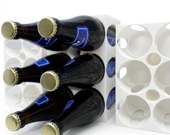 Op de afbeelding: Witte, modulaire wijnrek met verschillende donkere glazen flessen met gouden doppen en blauwe etiketten. Het rek is ontworpen met ronde openingen om de flessen veilig te houden. Een tweede, leeg rek is ook zichtbaar.