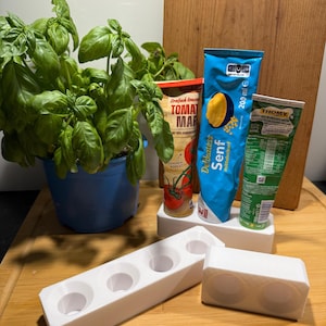 Könnte beinhalten: Ein blauer Topf mit frischem Basilikum neben einem Holzschneidebrett. Mehrere Lebensmittel werden gezeigt, darunter eine Tomatensaucendose, eine Senftube und eine grüne Tube. Weiße Halter mit kreisförmigen Öffnungen befinden sich im Vordergrund.