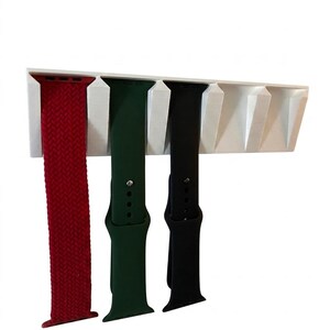 Könnte beinhalten: Ein weißer Wandhalter mit mehreren Schlitzen, an dem drei Uhrenarmbänder hängen. Die Bänder bestehen aus einem roten geflochtenen Armband, einem dunkelgrünen Silikonarmband und einem schwarzen Silikonarmband, die alle vertikal am Halter hängen.