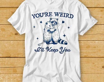 May include: White t-shirt with a blue graphic of a raccoon and opossum hugging. The text "You're Weird I'll Keep You" is printed in blue below the graphic.