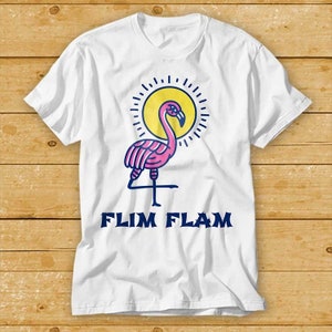 May include: White t-shirt featuring a pink flamingo illustration in front of a yellow sun with rays. The text "FLIM FLAM" is printed in blue below the flamingo.