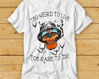 May include: White t-shirt featuring a skull wearing sunglasses and a hat, smoking a cigarette. The text "TOO WEIRD TO LIVE TOO RARE TO DIE" is printed above and below the skull. Black bat silhouettes are also included.