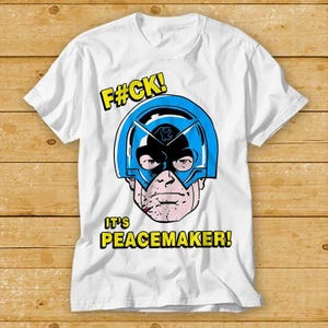 Könnte beinhalten: Weißes T-Shirt mit einer Comic-Illustration eines Charakters mit blauem Helm und Maske. Der Text "F#CK!" und "IT'S PEACEMAKER!" sind in Gelb gedruckt.