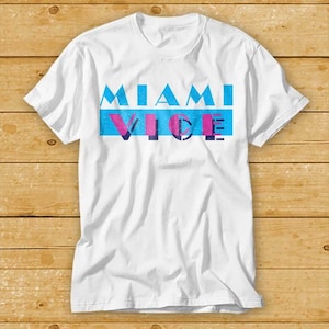 Può includere: Maglietta bianca con una stampa grafica blu e rosa che dice "Miami Vice" in stile retrò.