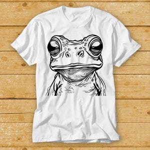以下が含まれることがあります： カエルの顔の白黒イラストが描かれた白いTシャツです。