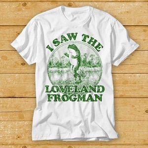 Puede incluir: Camiseta blanca con un gráfico verde de una rana en un estanque y el texto "I saw the Loveland Frogman".