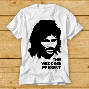 Könnte beinhalten: Weißes T-Shirt mit einer schwarzen Silhouette eines Männergesichts, langen Haaren und Bart. Der Text "THE WEDDING PRESENT" ist darunter in fetter Schrift gedruckt. Das T-Shirt wird auf einer Holzoberfläche präsentiert.