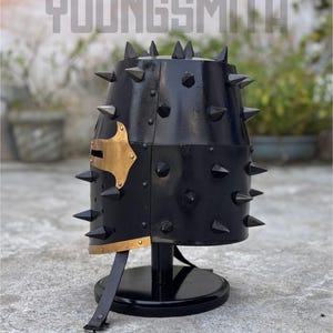 May include: Black metal helmet with numerous conical spikes. The helmet has a gold-colored accent on the side and is mounted on a black stand. The word "YOUNGSMITH" is visible at the top.