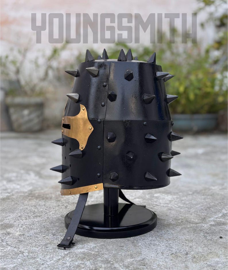 May include: Black metal helmet with numerous conical spikes. The helmet has a gold-colored accent on the side and is mounted on a black stand. The word "YOUNGSMITH" is visible at the top.