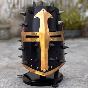 May include: A black medieval helmet with gold accents and spikes. The helmet features a gold cross design on the front and is mounted on a black stand. The word "YOUNGSMITH" is visible at the top.