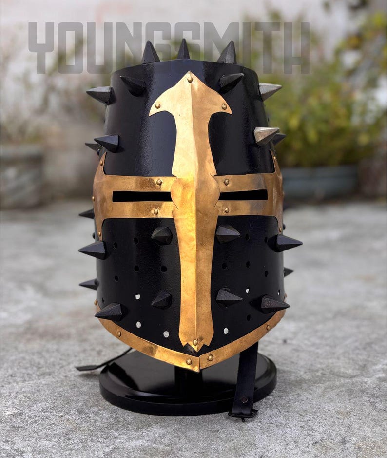 May include: A black medieval helmet with gold accents and spikes. The helmet features a gold cross design on the front and is mounted on a black stand. The word "YOUNGSMITH" is visible at the top.