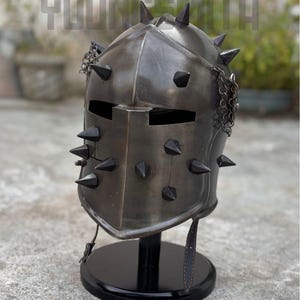 May include: A metallic, spiked helmet with a dark finish, displayed on a black stand. The helmet features multiple black spikes and a chain detail. The text "YOUNG SMITH" is visible at the top.