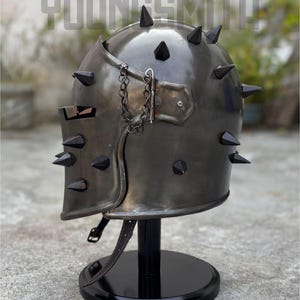 May include: A dark metal helmet with black spikes and a chain detail. The helmet is displayed on a black stand. The word "YOUNGSMITH" is visible in the background.